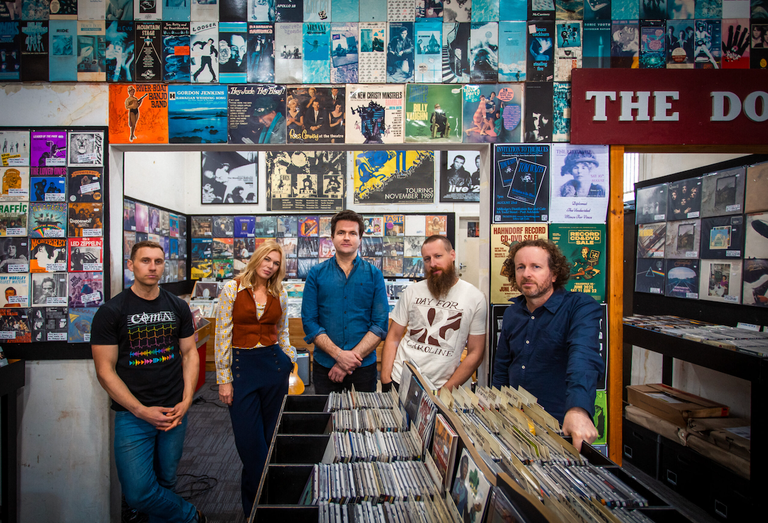 Band photo of Jen Lush & The Field standing inside a retro vinyl record shop surrounded by wall-to-wall album covers and record bins.